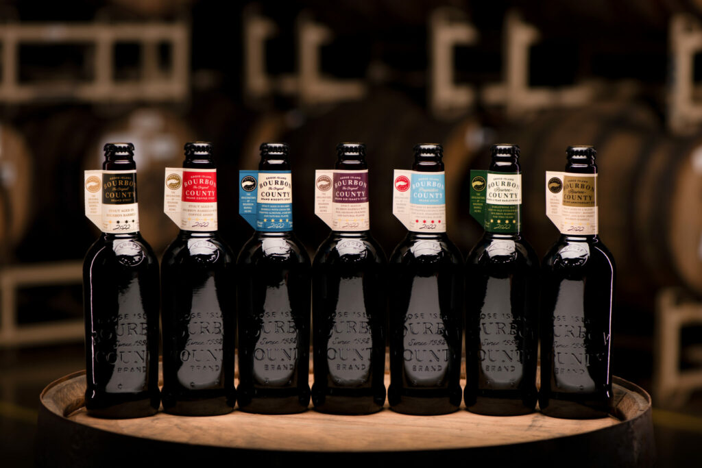 The 2022 Bourbon County variants. Photo is of seven black bottles sitting on top of a bourbon barrel, with other barrels in the background. 